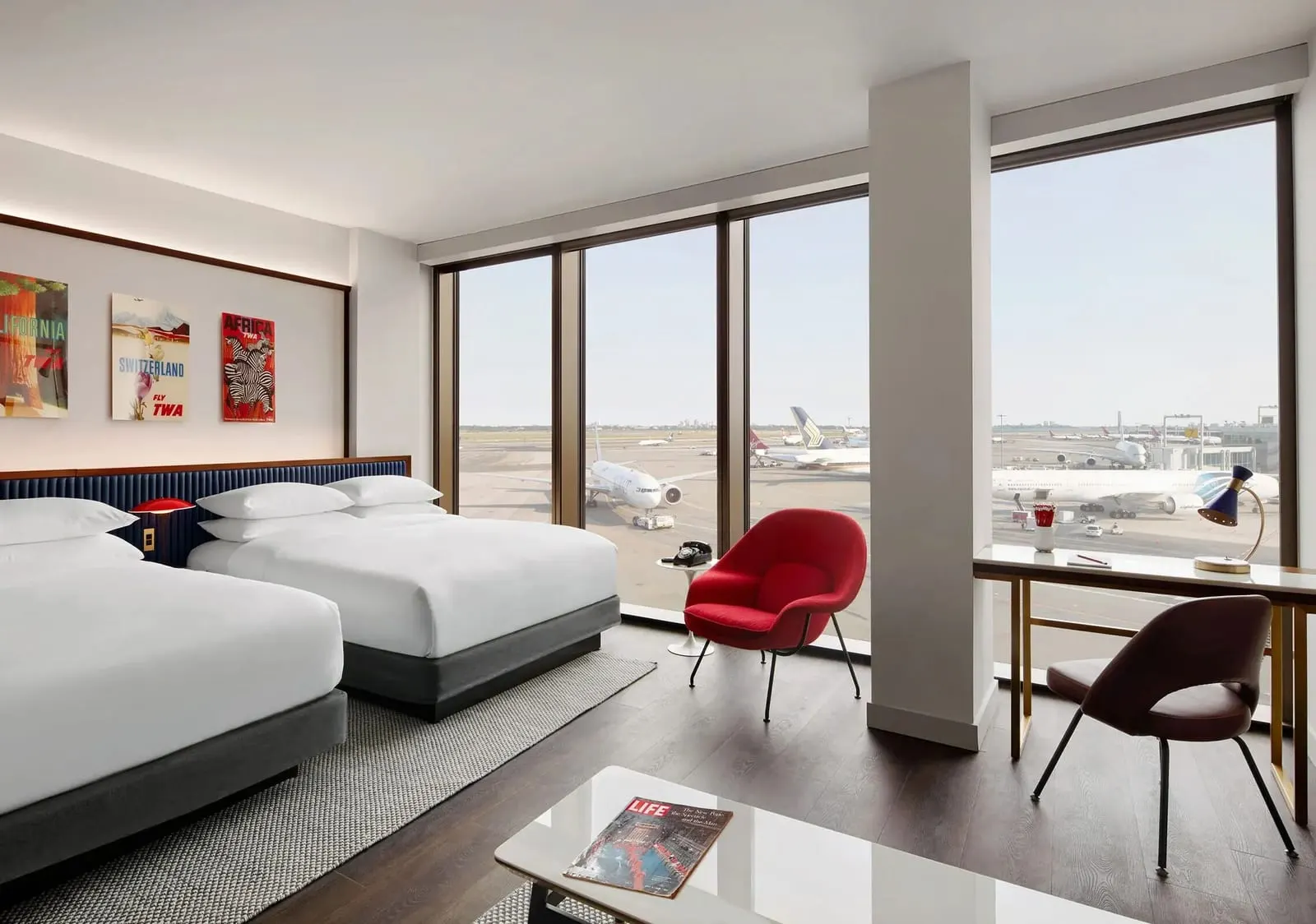 Executive Double Queen Suite With Runway View at TWA Hotel: two queen beds, a red armchair and a desk beside corner windows that look over airliners on the apron