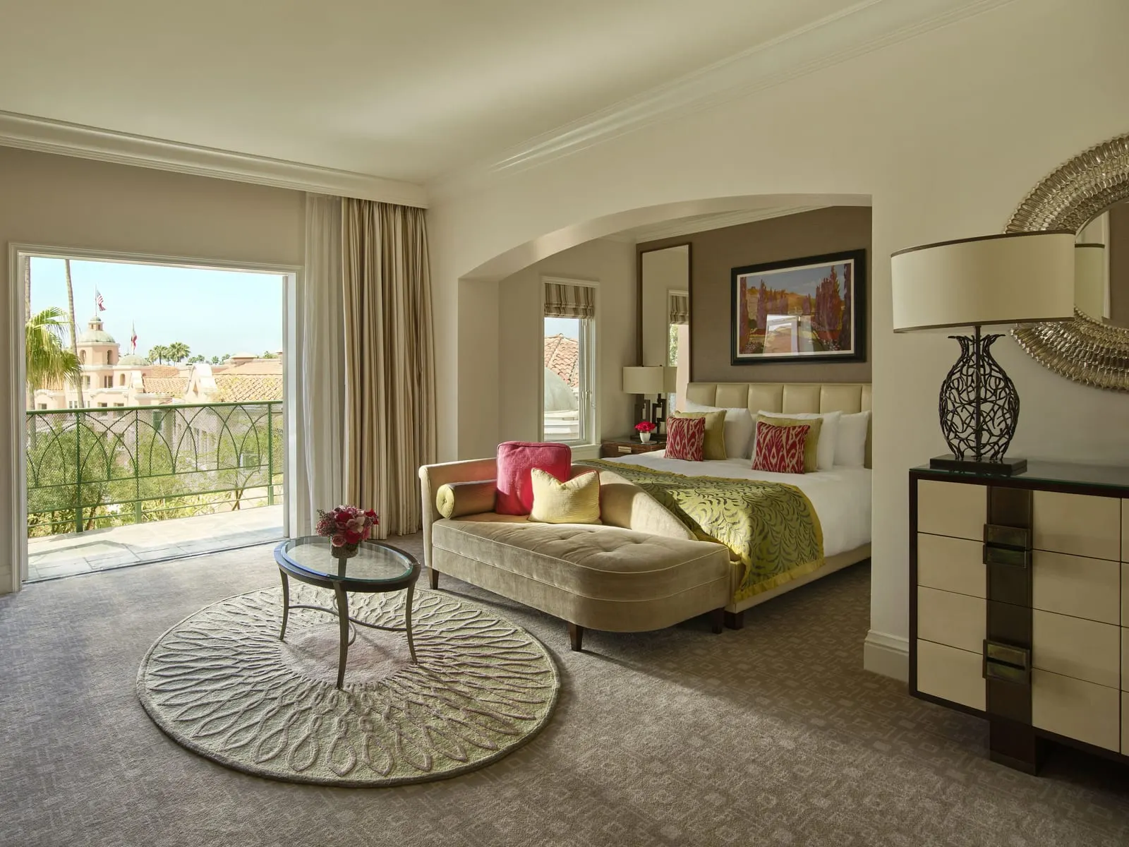 Deluxe Room with Balcony at The Beverly Hills Hotel: a bed in an arched alcove with a tan chaise at its foot and a round rug, and an open doorway to a balcony with a green iron railing