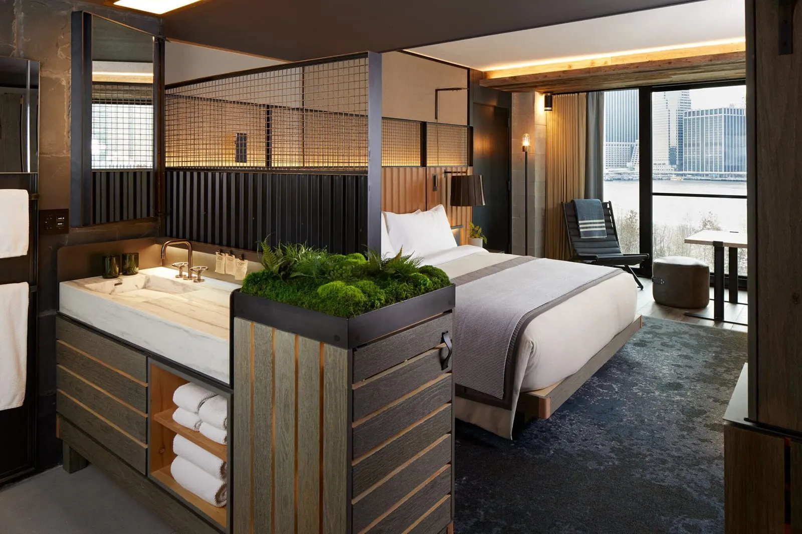 Liberty King im 1 Hotel Brooklyn Bridge: ein Kingsize-Bett und ein offenes Marmorwaschbecken hinter einem Pflanzkasten mit Moos, mit einem raumhohen Fenster zum East River und zur Skyline von Lower Manhattan