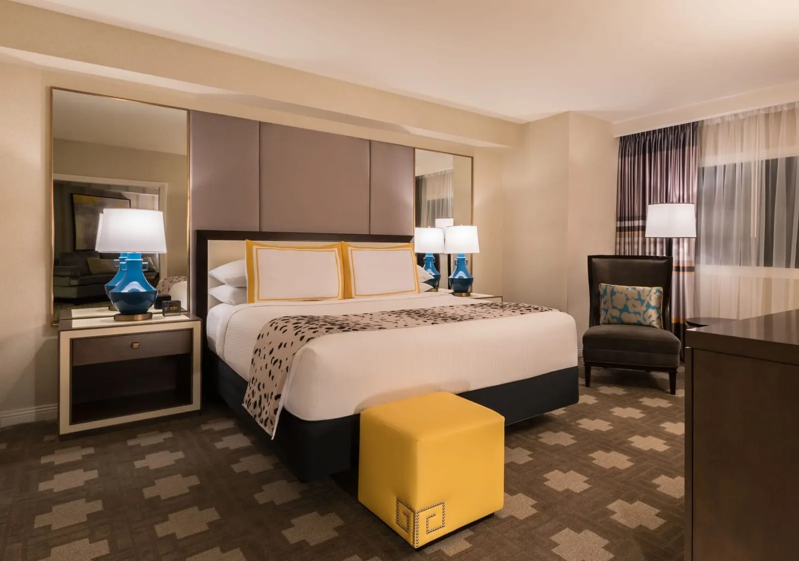 Julius Studio Suite im Caesars Palace: ein Kingsize-Bett mit einem gelben Polsterhocker, blauen Lampen und einem dunklen Sessel am Fenster