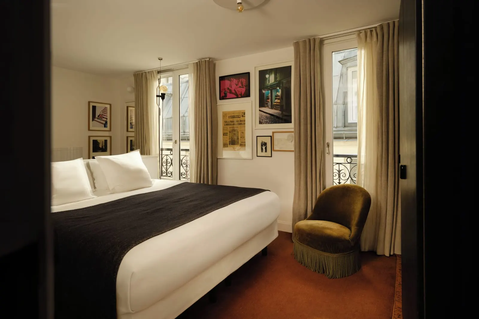 Pigalle 22 at Le Pigalle: a double bed with a dark throw, a fringed velvet armchair and two French windows with wrought-iron railings, under framed photographs and an old newspaper front page