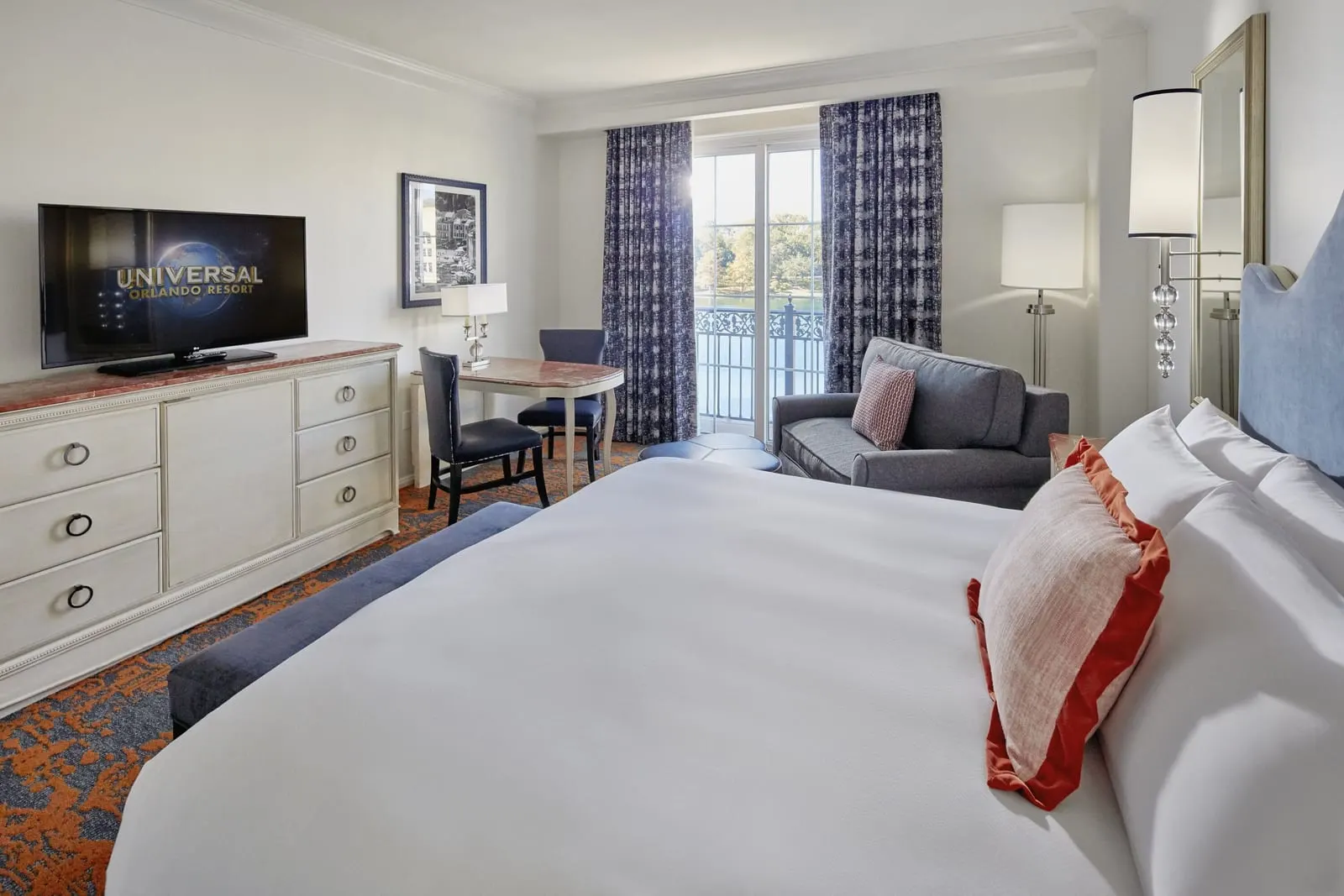 Bay View King Room at Loews Portofino Bay Hotel: a king bed facing a dresser with a television, with a writing desk, two chairs and an armchair beside tall curtained windows that look out over the water