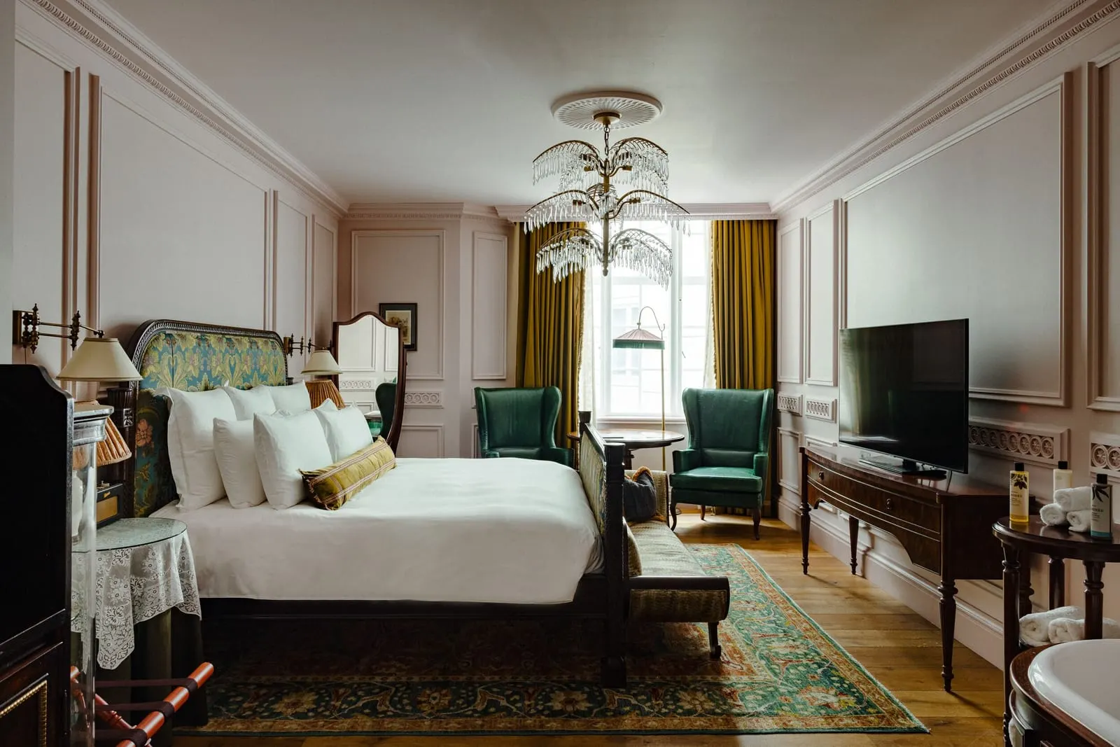 Large room at The Ned: the bed, green wingback chairs, a glass leaf chandelier and a television