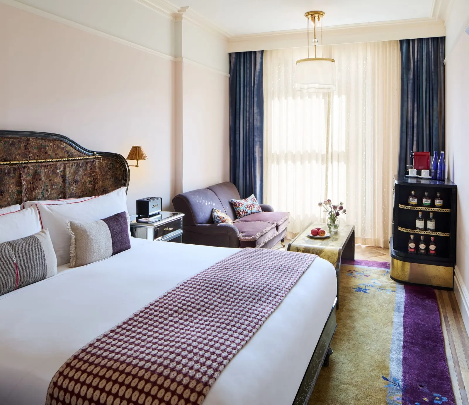 Grand Chelsea at Hotel Chelsea: a bed with a velvet headboard, a purple sofa and a low coffee table in front of a tall curtained window, and a black drinks cabinet in the corner