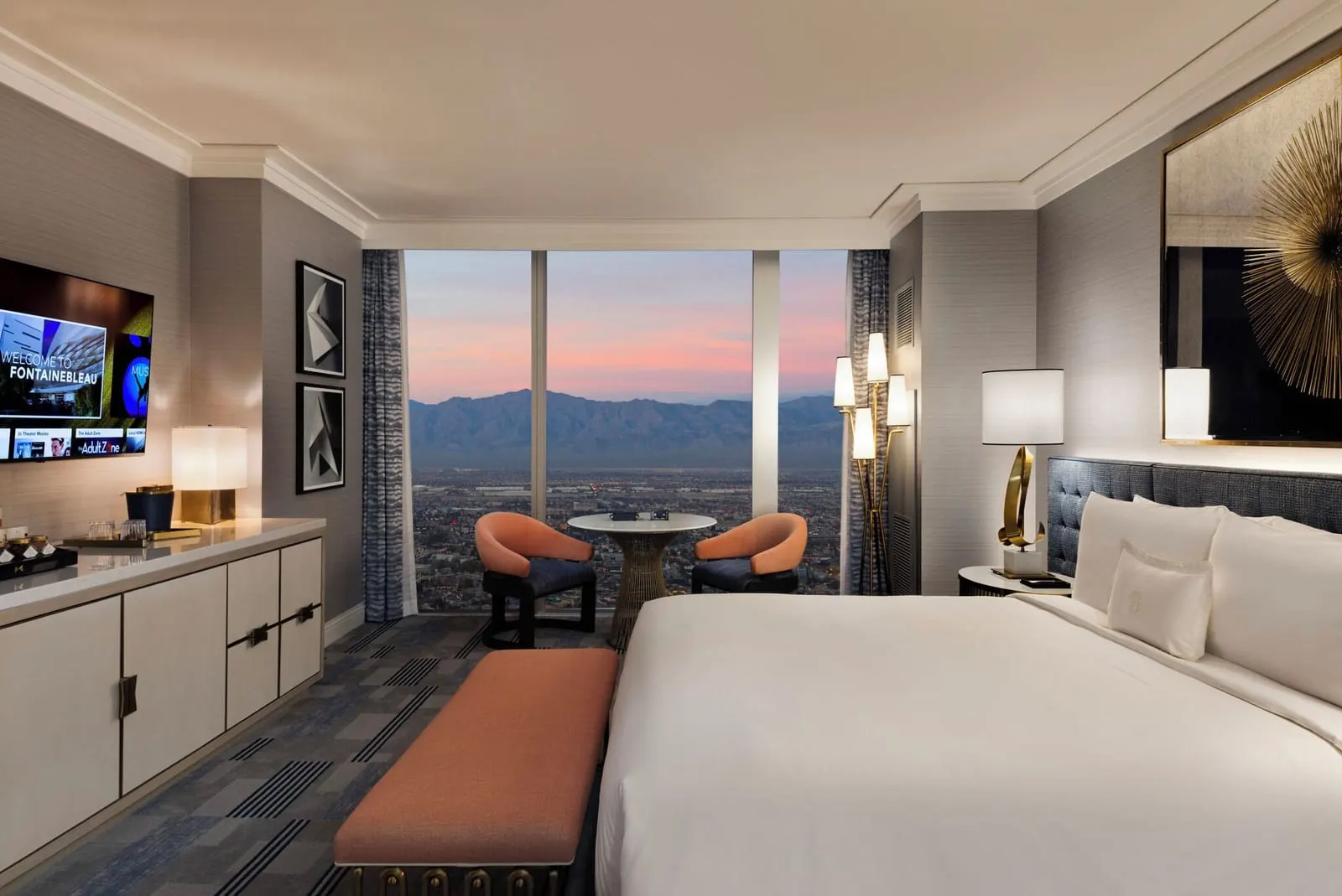 Bleau King Mountain Great View at Fontainebleau Las Vegas: a king bed, a long cabinet under a wall television and two chairs at a round table by floor-to-ceiling windows onto the city and the mountains at dusk