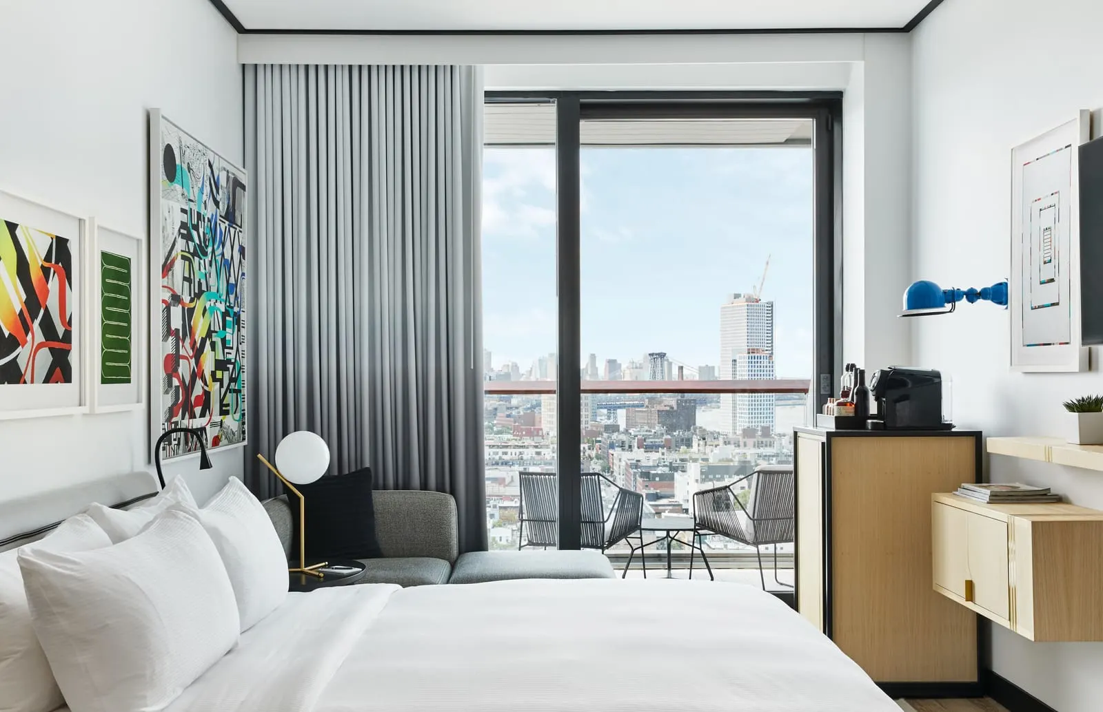 King with Balcony at The William Vale: a bed with white linen facing a glass balcony door, with two chairs on the balcony and rooftops beyond, a coffee station and colourful framed prints