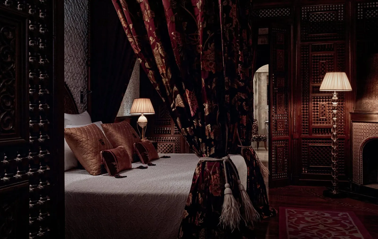 Prestige Riad at Royal Mansour Marrakech: a bed hung with heavy patterned curtains, carved wooden screens and a tiled fireplace in a lamplit bedroom