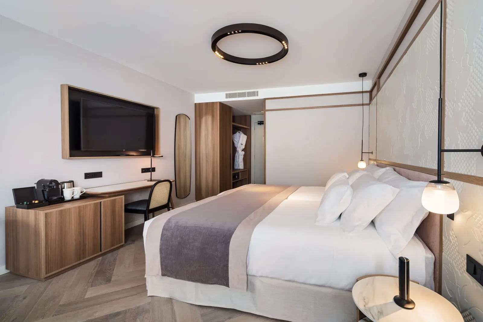 Junior Suite at Barceló Funchal Oldtown: the bed against a carved white headboard panel, facing a wall television, a desk and an open wardrobe with bathrobes