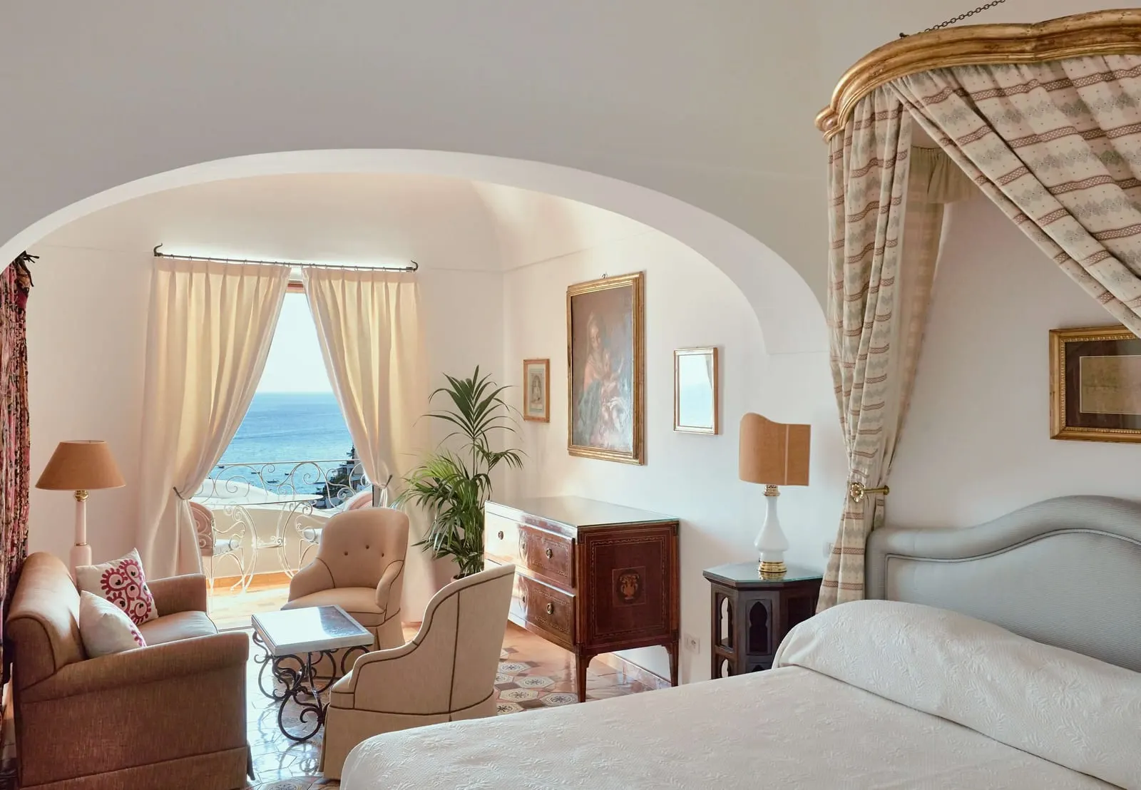 Superior Sea View at Le Sirenuse: a canopied bed beside a sitting area of armchairs, with a curtained window onto a balcony over the sea