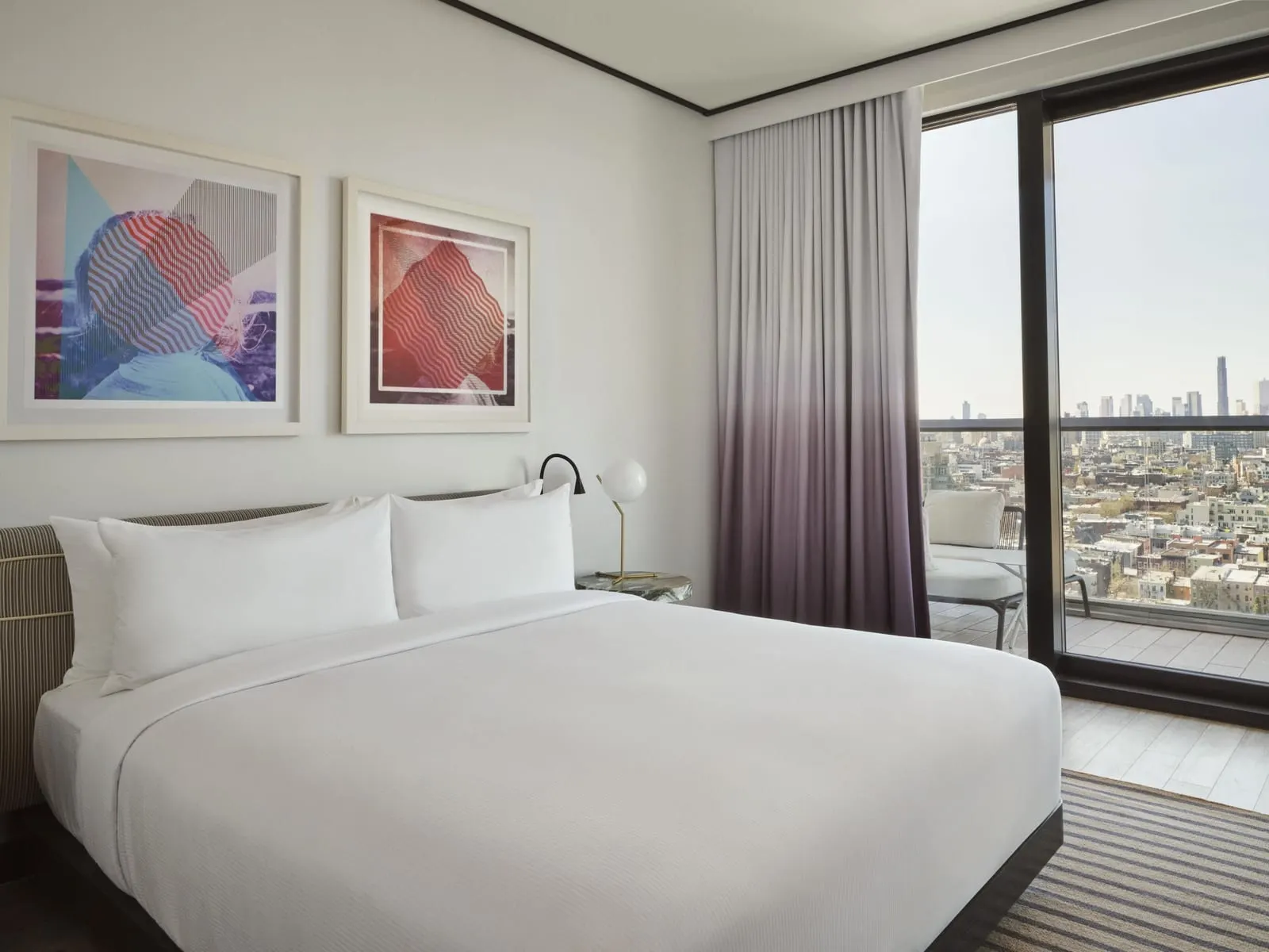 King One Bedroom Suite with Balcony at The William Vale: a bed with white linen under two framed abstract prints, a bedside lamp with a white globe shade, and a floor-to-ceiling window over low-rise rooftops