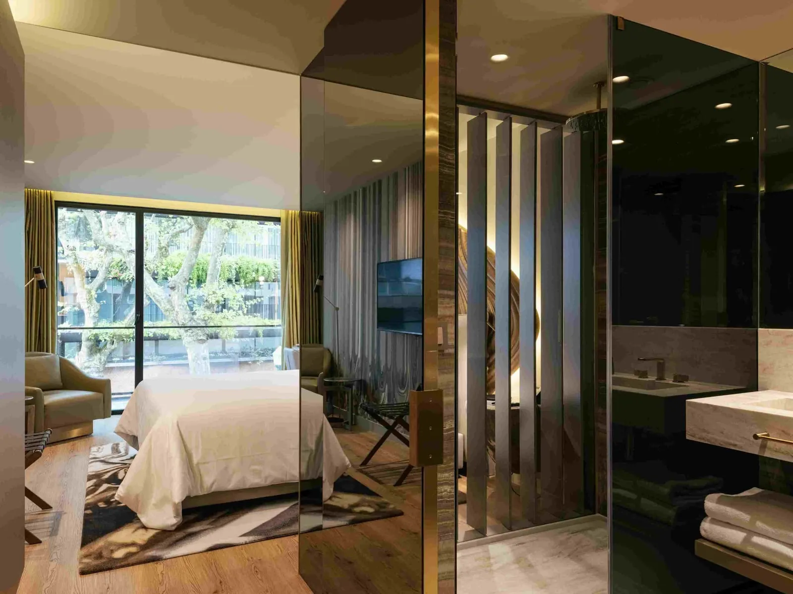 Onice Room at Vila Foz: the bed, an armchair and a window onto trees, with the glass-walled bathroom on the right