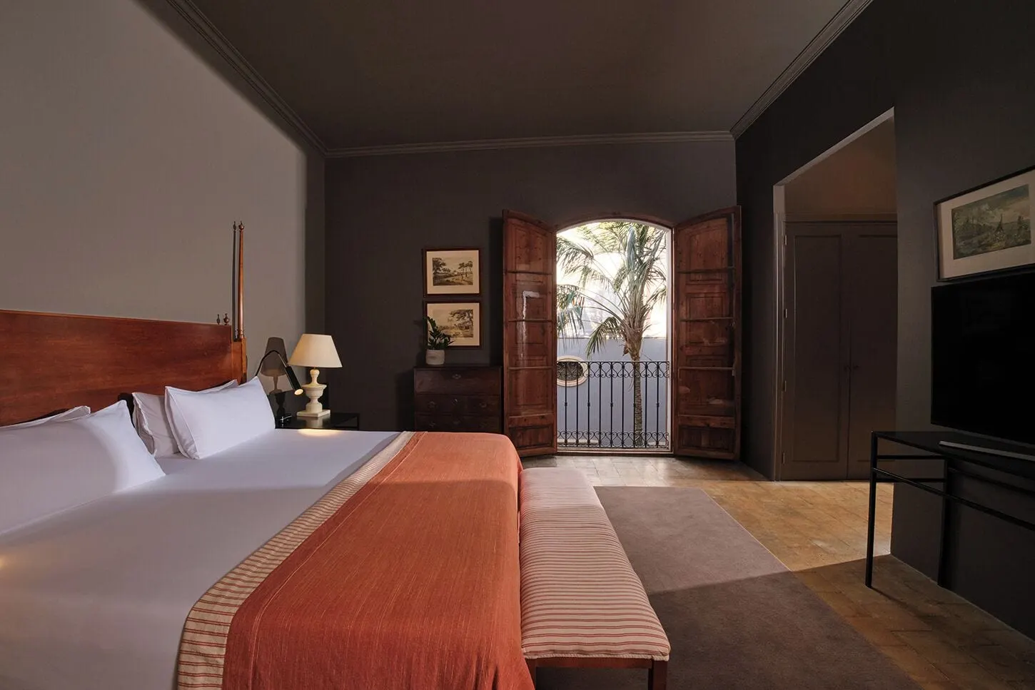 Premium im Can Cera: ein Bett mit einer orangefarbenen Tagesdecke und einer gestreiften Bank, ein Fernseher und Bogentüren mit Fensterläden, die sich auf einen Balkon mit einer Palme dahinter öffnen