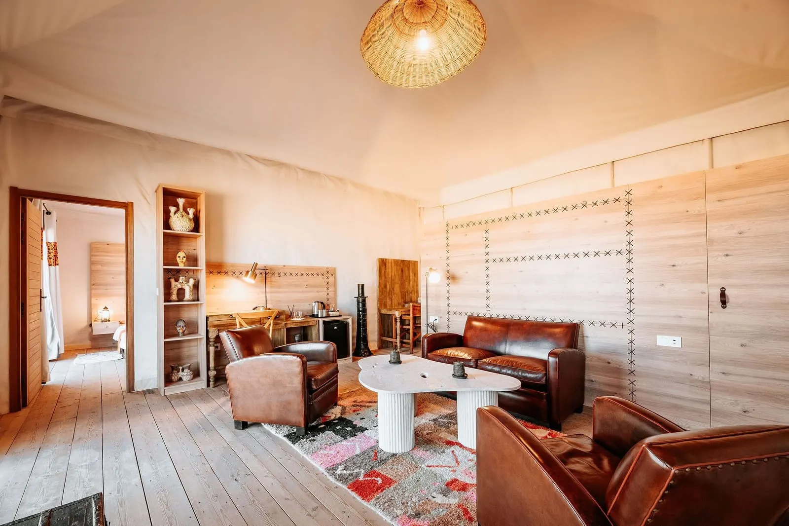 Suite n°19 at Inara Camp: a lounge with leather armchairs, a leather sofa and white round tables on a patterned rug, with a doorway to the next room.