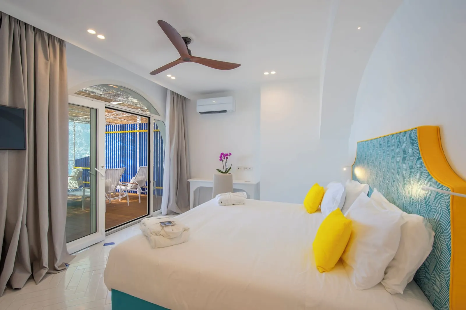 Deluxe Room with Jacuzzi and sea view terrace all'Hotel Tramonto d'Oro: un letto matrimoniale con testiera turchese e gialla sotto un ventilatore a soffitto, con porte di vetro su una terrazza con pavimento in legno.