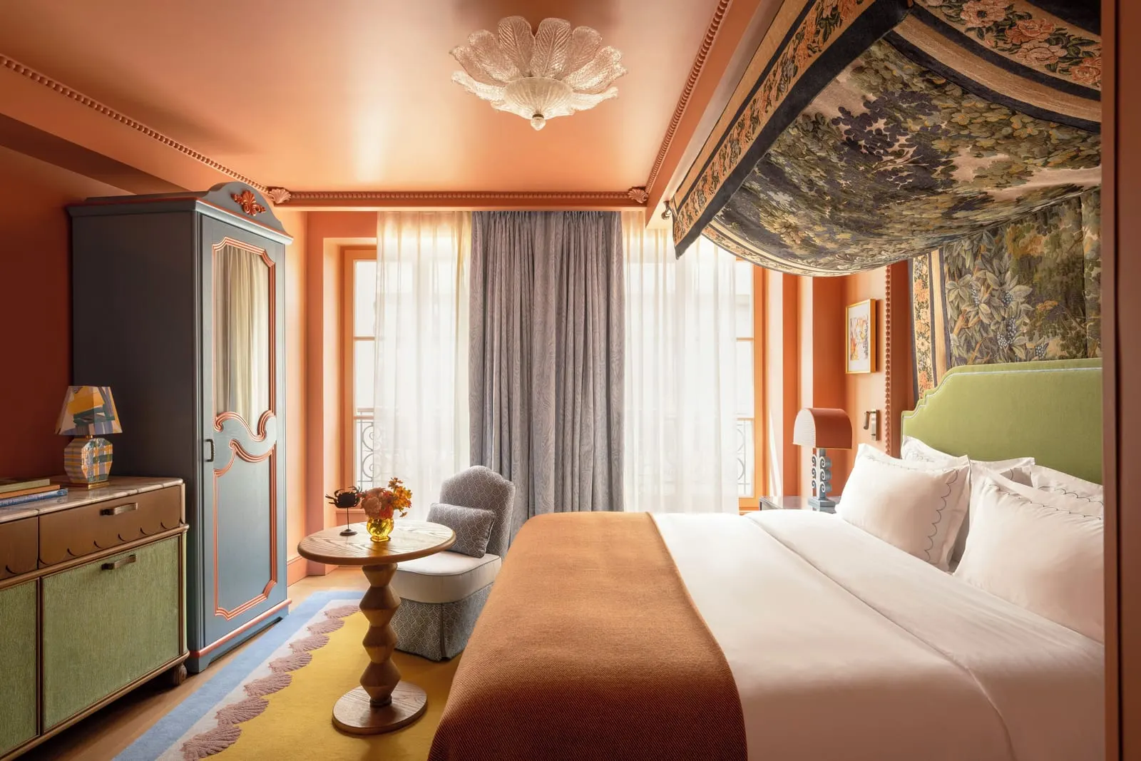 Superior Room at Le Grand Mazarin: a bed with a green velvet headboard under a tapestry canopy, terracotta walls, a painted wardrobe and an armchair by tall windows with grey curtains