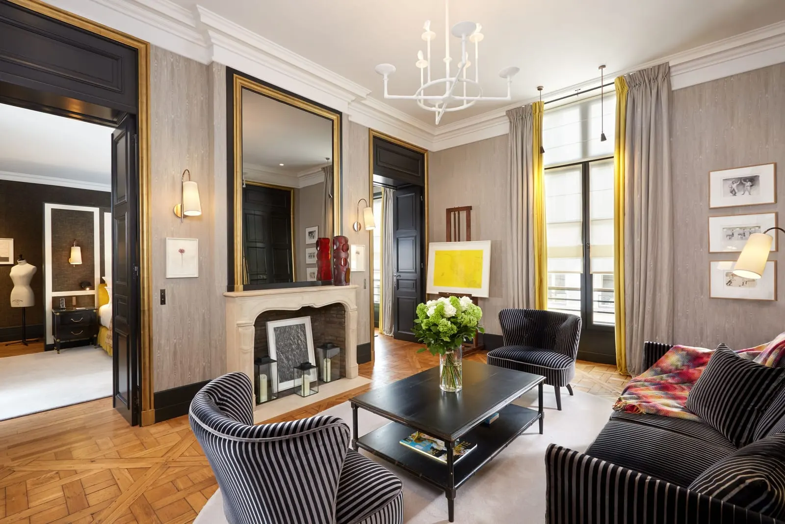 Queen's Suite at Le Pavillon de la Reine: the living room, with a stone fireplace, a white chandelier, gilt-edged doorways, striped armchairs and Versailles parquet, and the bedroom through tall double doors