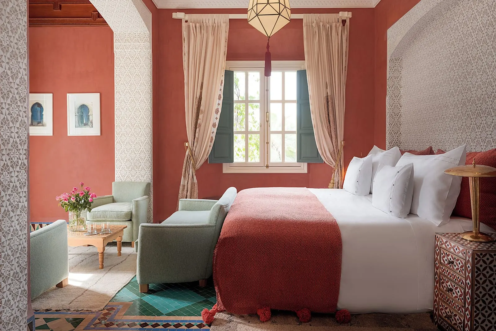 La Favorite at Dar Rhizlane: a king-size bed with a terracotta throw beside a shuttered window, and armchairs round a low table through the archway