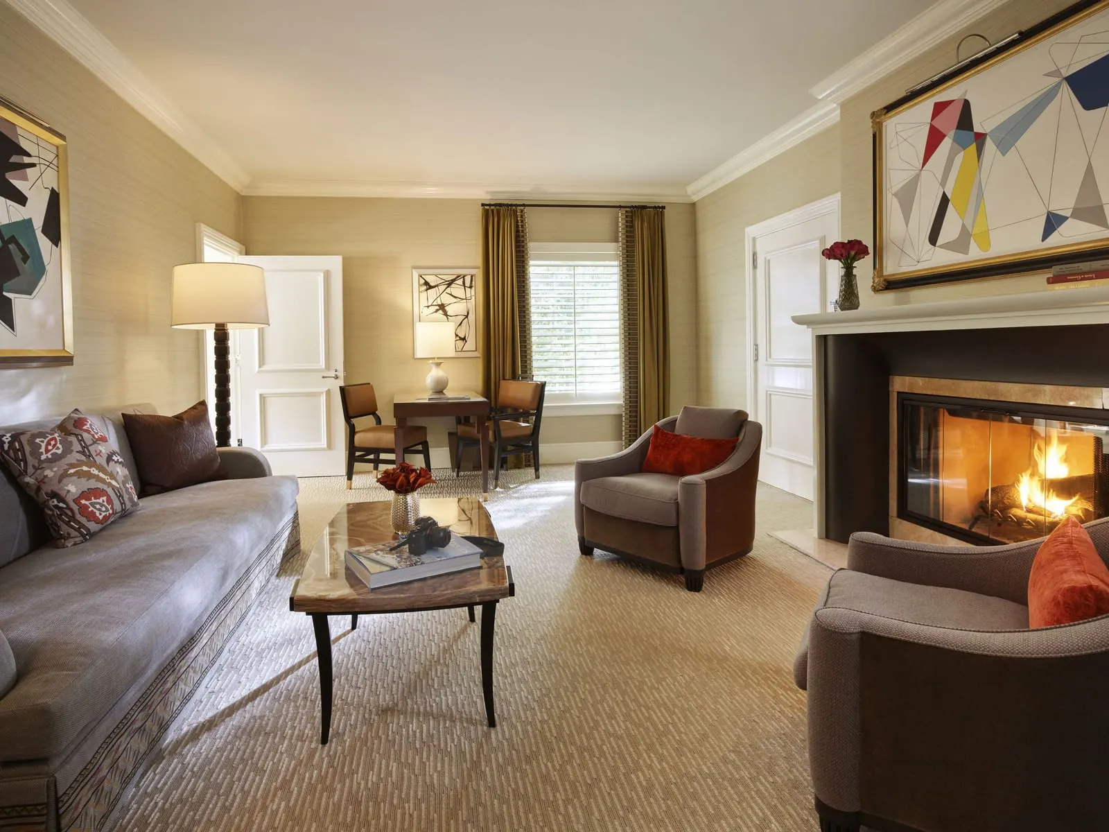 Beverly Hills Bungalow Suite at The Beverly Hills Hotel: a living room with a grey sofa, two armchairs and a stone-topped coffee table around a lit fireplace, and a small table with two chairs by a shuttered window