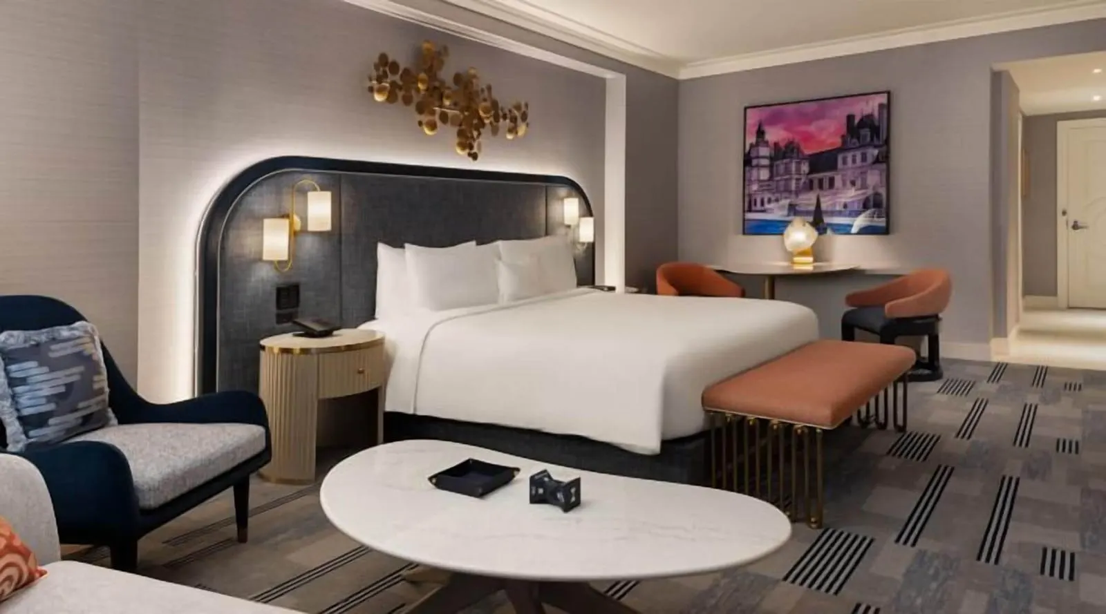 Gold King Pool View at Fontainebleau Las Vegas: a king bed against a curved navy headboard with gold wall lights and a gold disc sculpture above it, a peach bench, a navy armchair and an oval marble coffee table