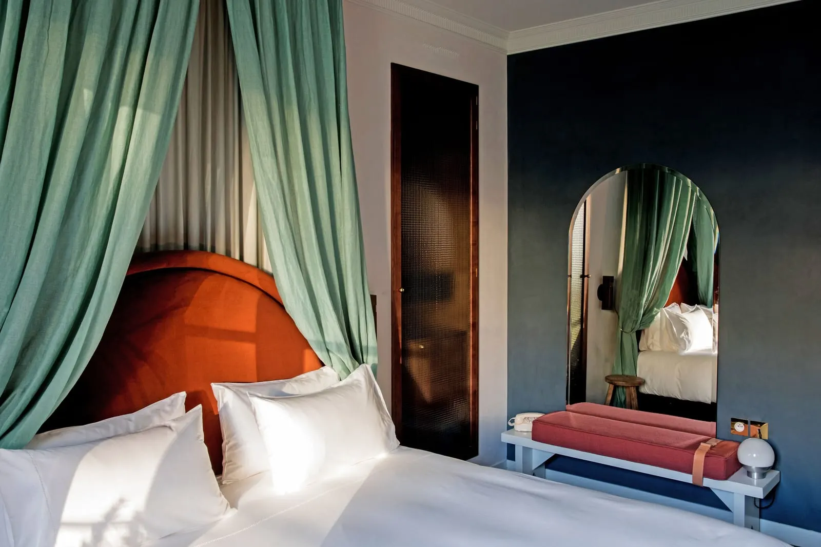 Uzes at Grands Boulevards Experimental: a bed under a sage green canopy with a rust velvet headboard, beside a dark blue wall with a tall arched mirror above a white bench with a coral cushion