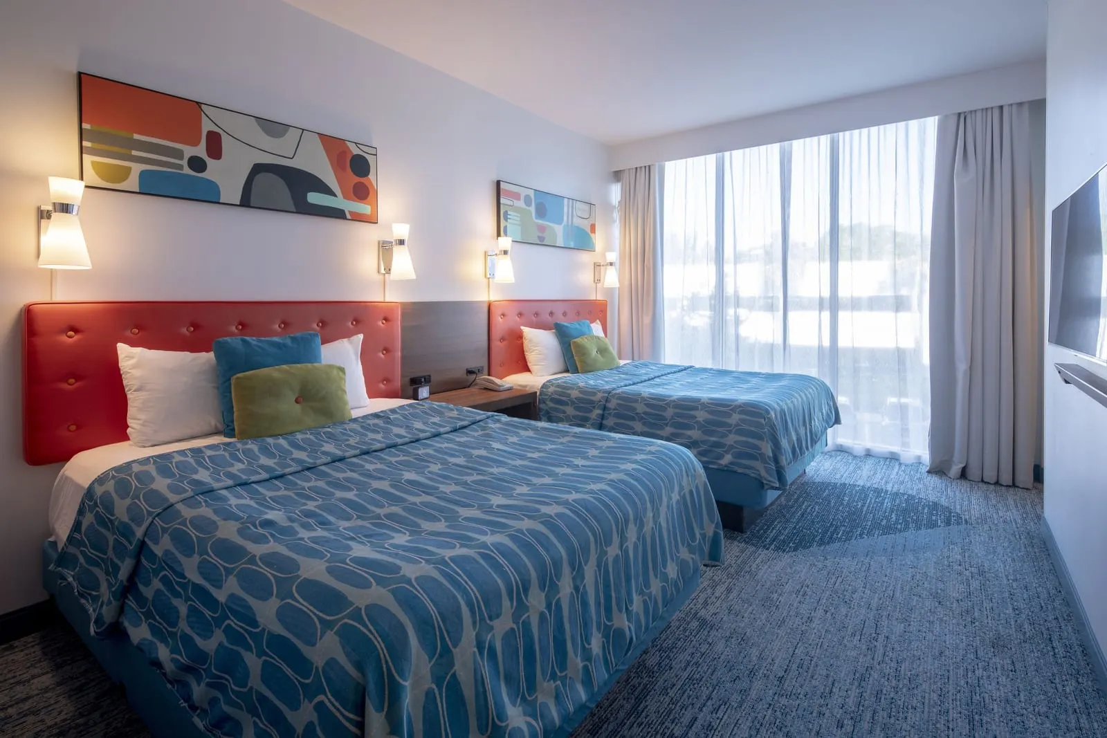 Family Suite Interior Entry at Universal Cabana Bay Beach Resort: two queen beds with orange headboards and blue patterned covers, a wall-mounted television and a wide window with sheer curtains