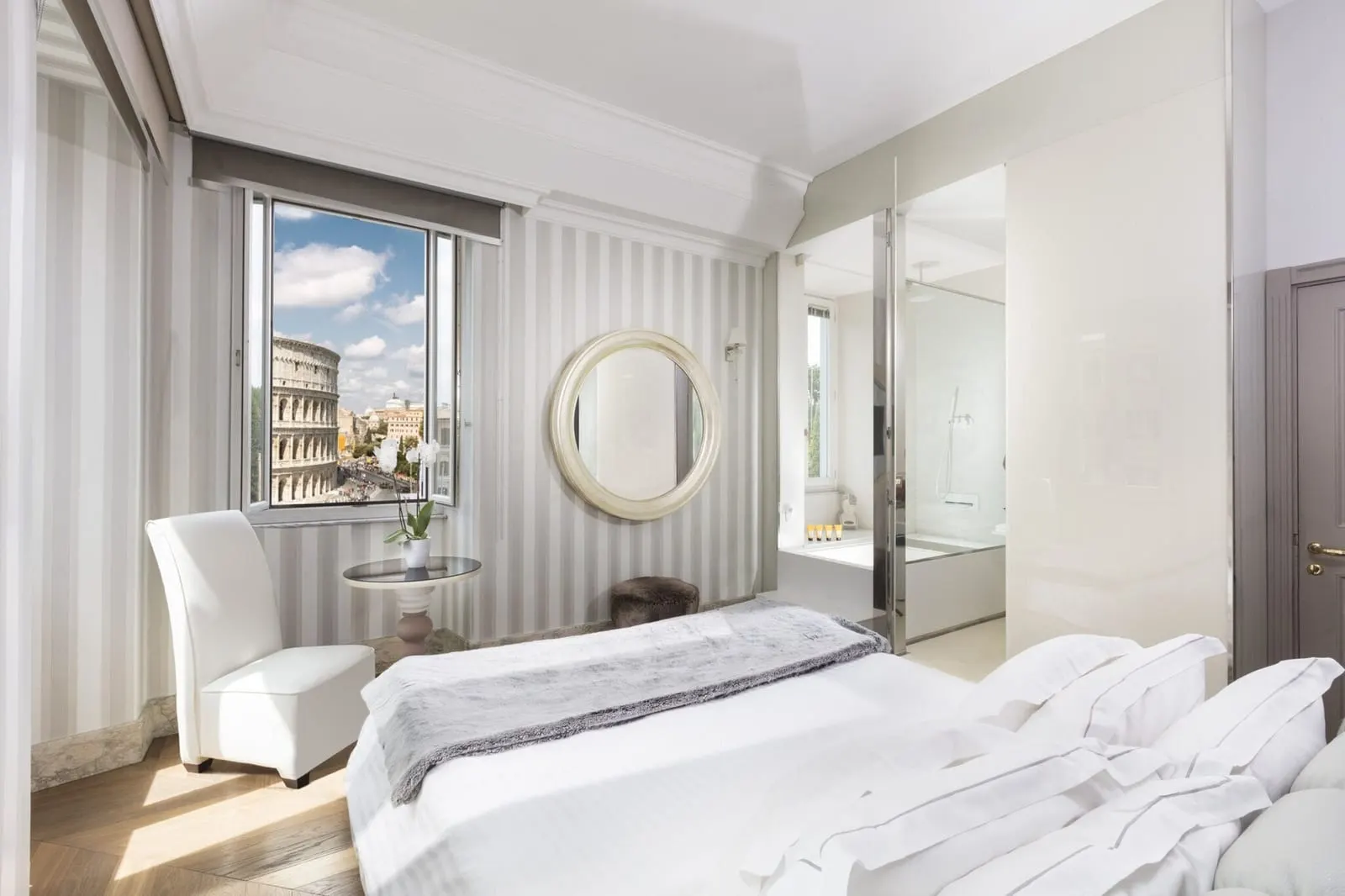 Master Room at Palazzo Manfredi: the bed, a white armchair and a round mirror, with the Colosseum in the window