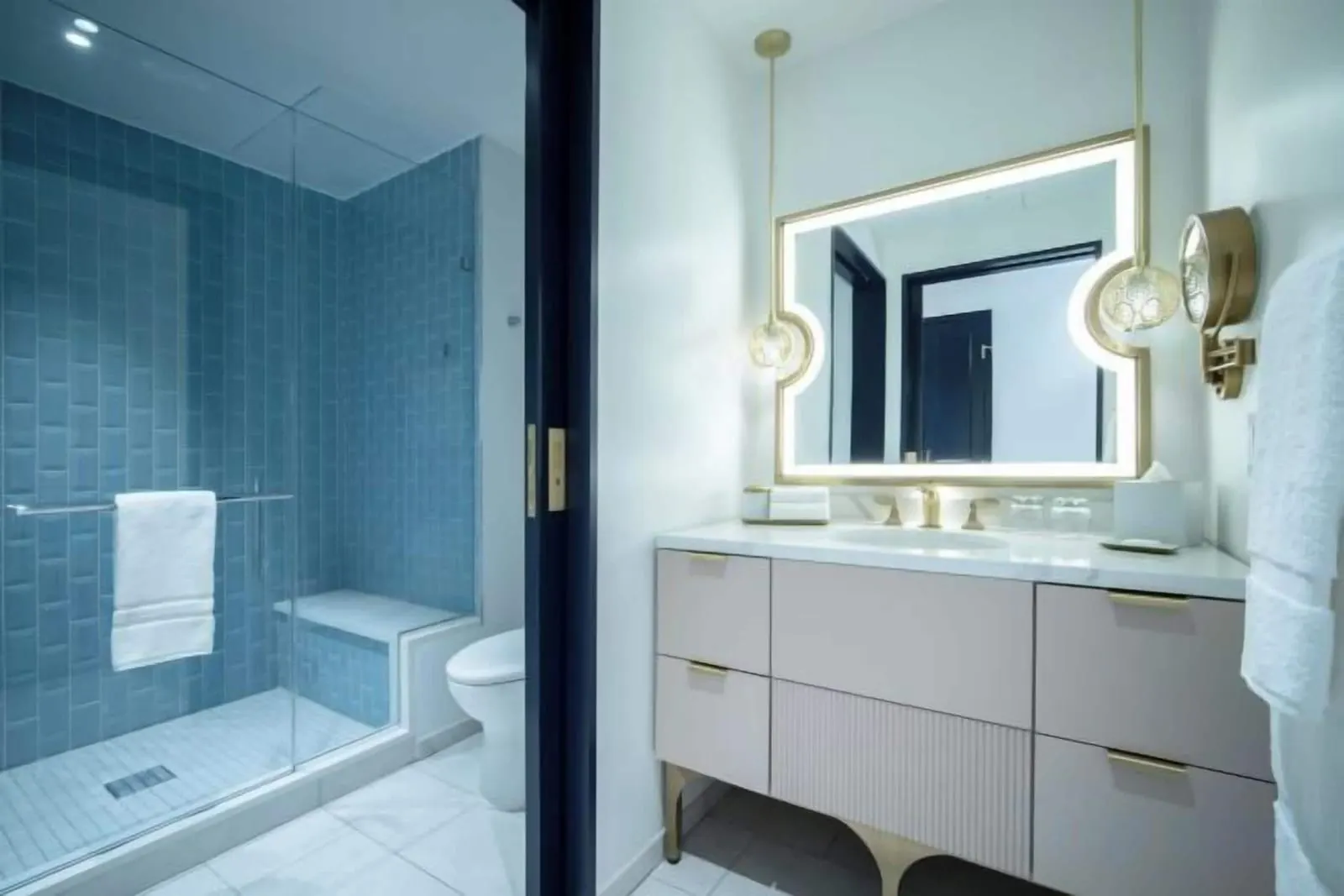Pool View 2 Queen Beds room at Universal Helios Grand Hotel: the bathroom, with a blue-tiled walk-in shower with a bench and a toilet behind a sliding door, and a separate vanity with a lit mirror
