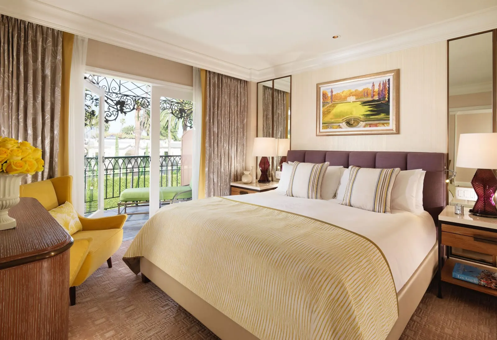 Junior Beverly Hills Suite at The Beverly Hills Hotel: a bed with a plum headboard and a yellow patterned throw, a yellow armchair, and French doors open to a balcony with a wrought-iron railing and a lounger