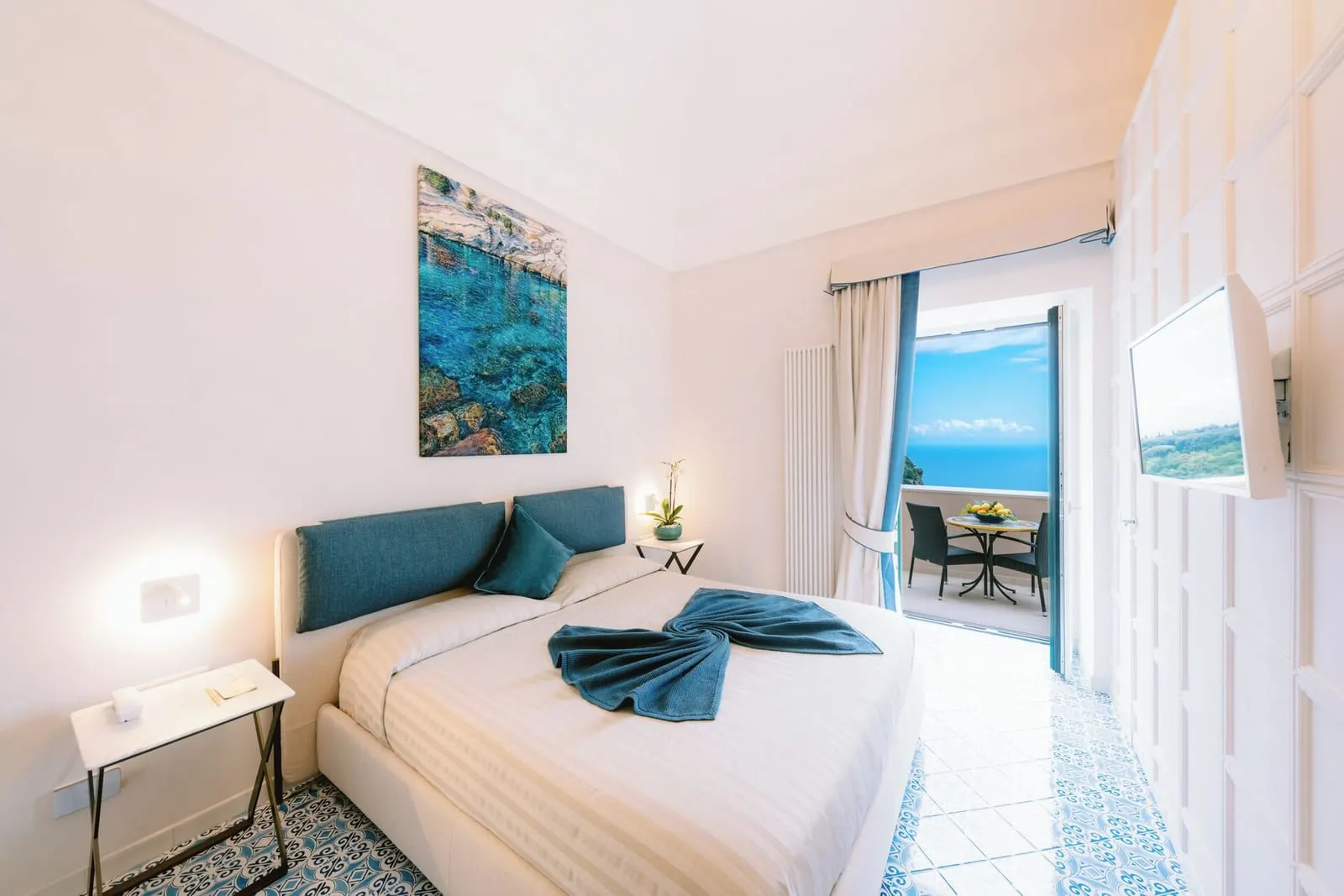 Grand Junior Suite MELISSA at Palazzo Pascal: a double bed with a teal headboard and a glass door open onto a terrace with chairs over the sea