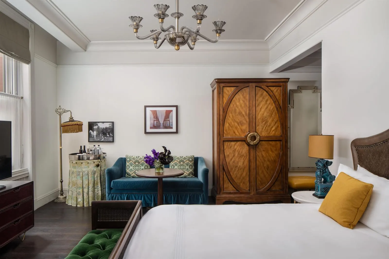 Studio Suite at The Beekman: a bed in the foreground, a blue velvet sofa with a round table, a tall wooden armoire and a skirted drinks cart under a chandelier