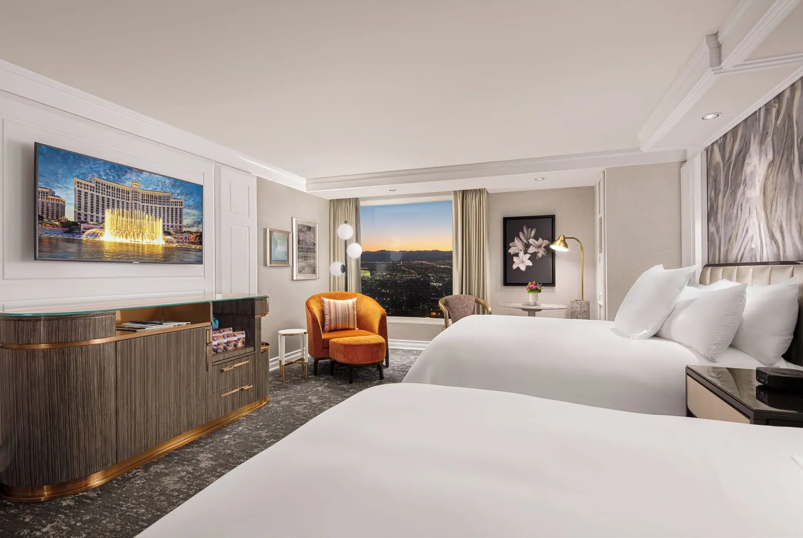 Premier Two Queen at Bellagio: two queen beds, an orange armchair and footstool by the window, and a long wooden cabinet under a wall-mounted television