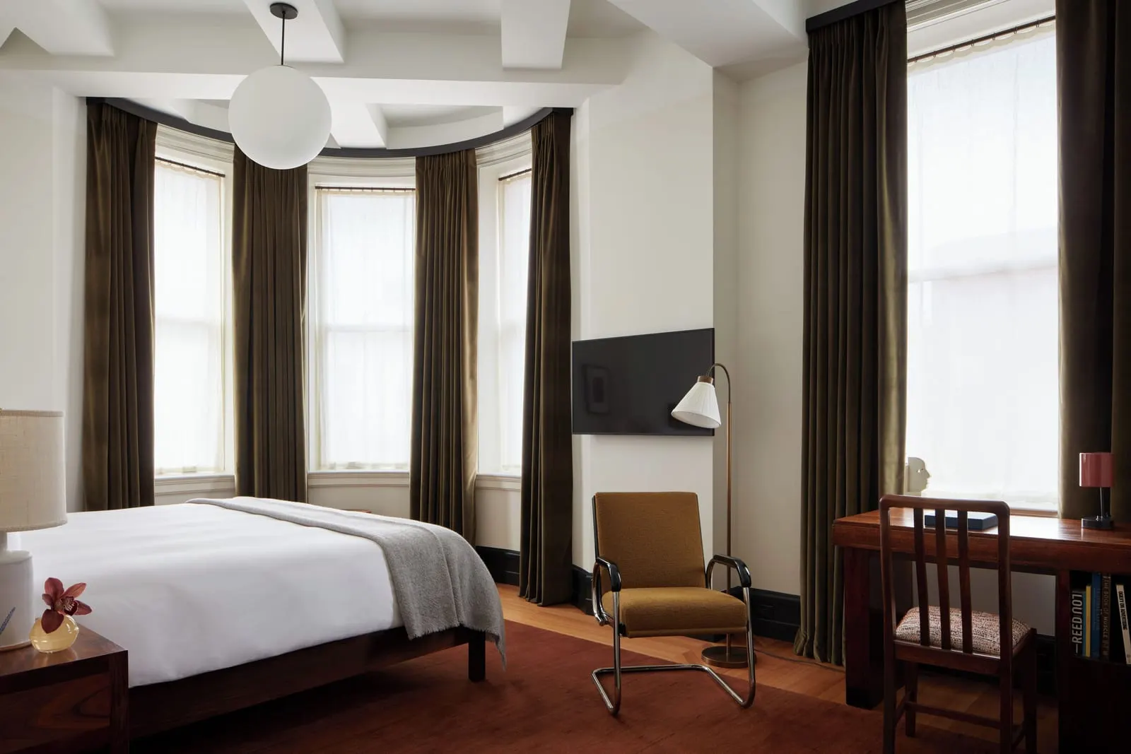Corner King at Nine Orchard: a bed with a grey wool throw beside a curve of three tall windows in the rounded corner, with a yellow armchair, a wall-mounted television and a wooden desk