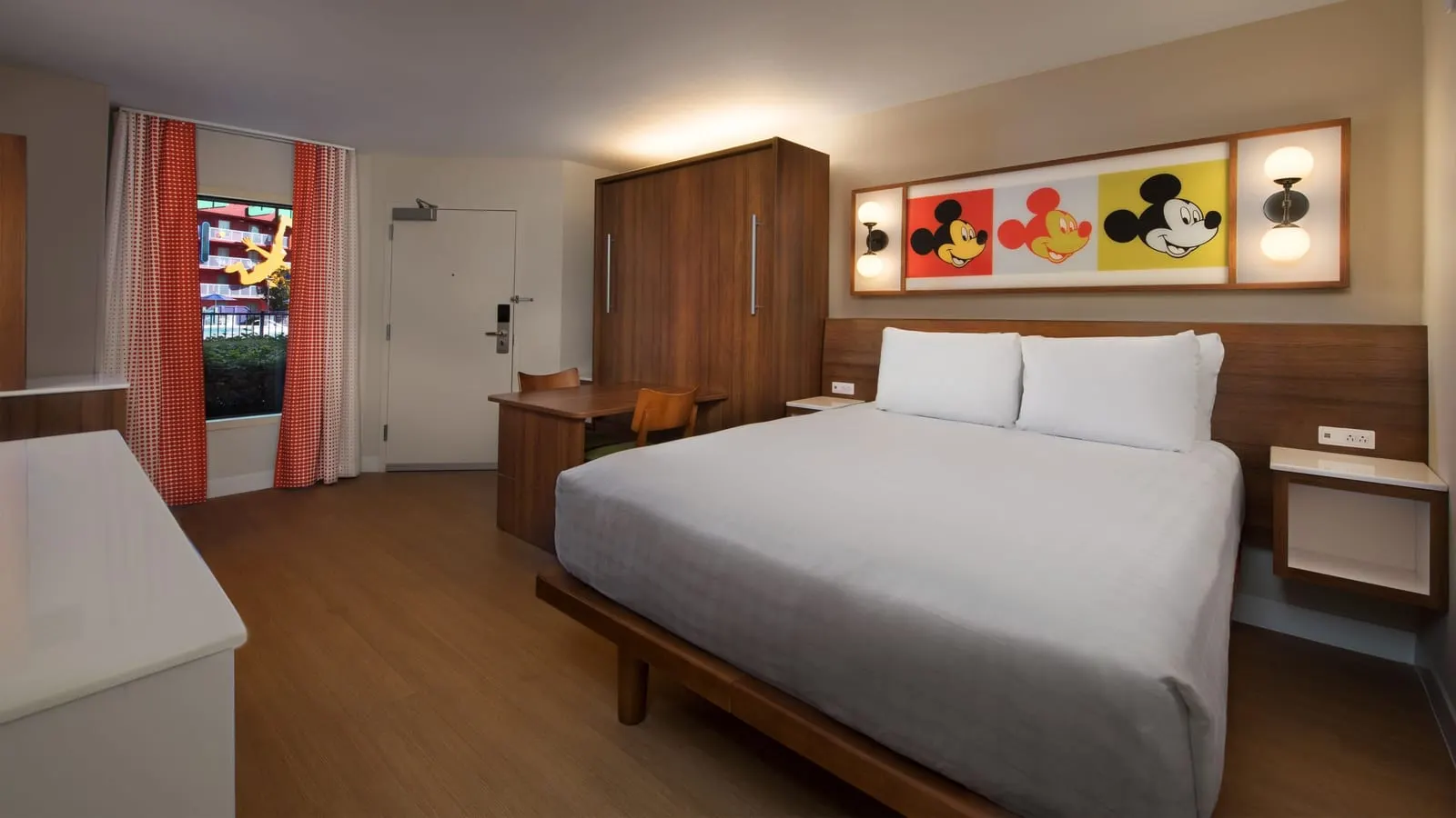 Preferred Pool View room at Disney's Pop Century Resort: a queen bed under a framed Mickey Mouse print, the tall wooden cabinet of the fold-down second bed with a table and two chairs, and a curtained window beside the door
