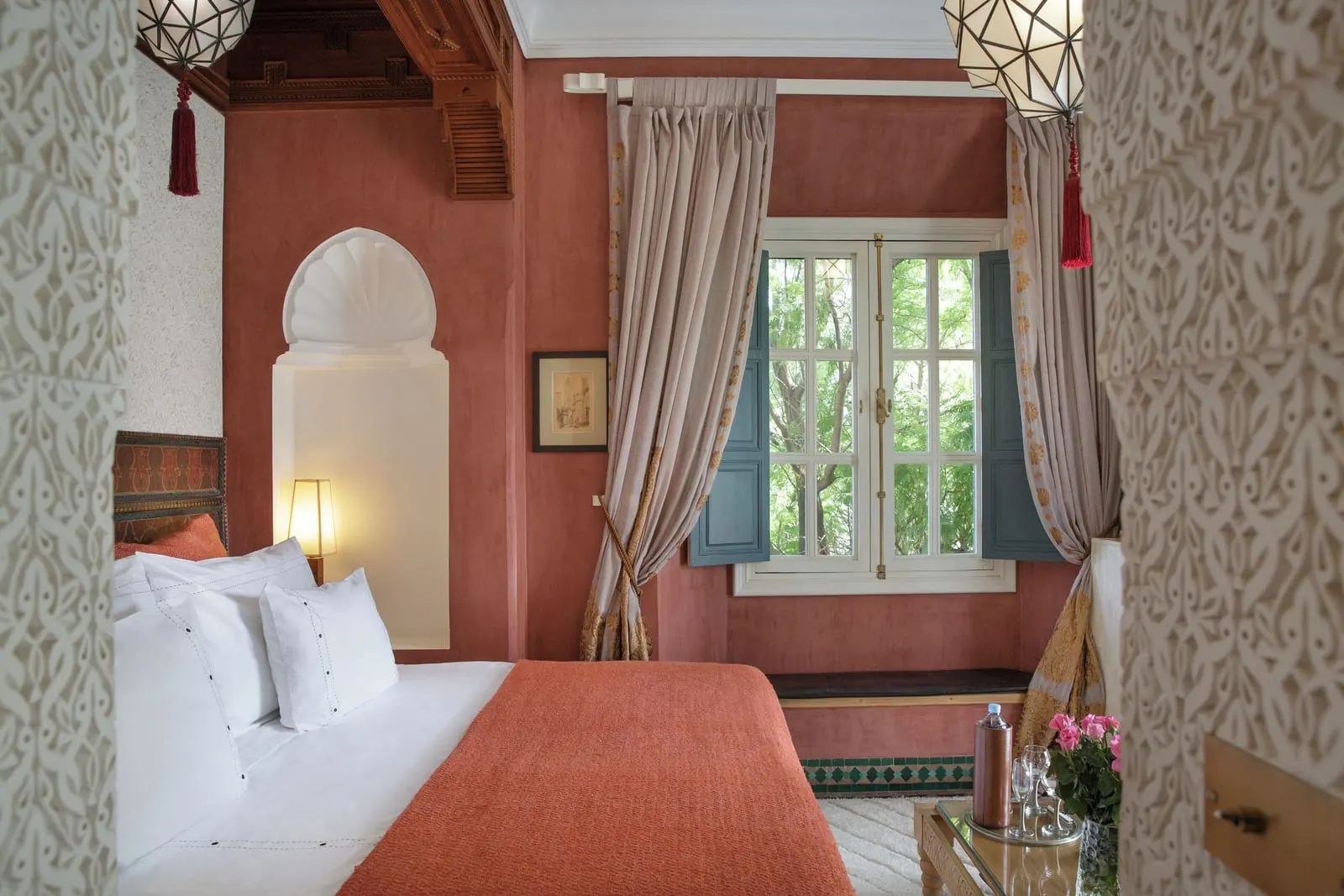 La Favorite at Dar Rhizlane: the bed beside a shell-carved alcove with a lamp, red walls and a window framed by long curtains