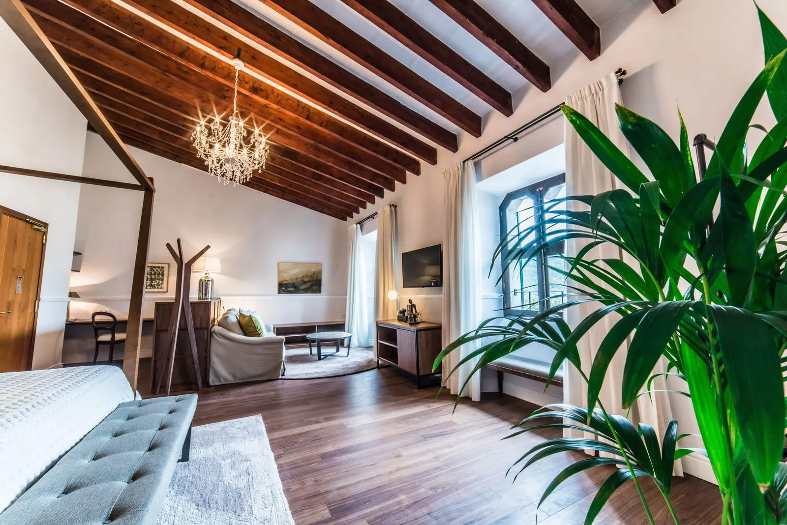 Luxury Suite at L'Avenida Hotel: the sitting area beyond the bed, with a sofa, a desk and a tall window under a beamed ceiling and chandelier