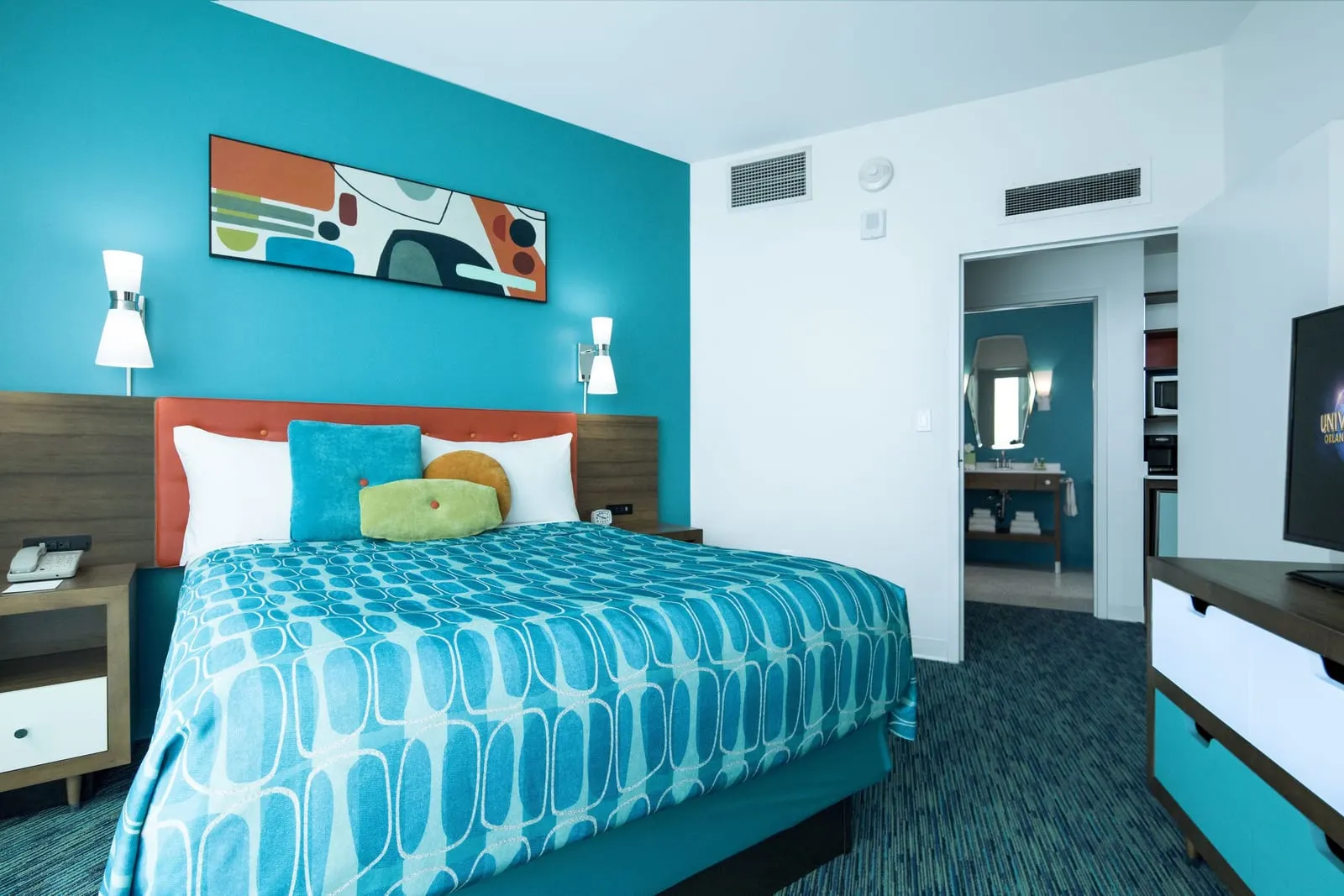 Volcano Bay View 2-Bedroom Suite at Universal Cabana Bay Beach Resort: the queen bedroom, with an orange headboard against a turquoise wall, a dresser with a television and a doorway through to a washbasin and the kitchenette with a microwave