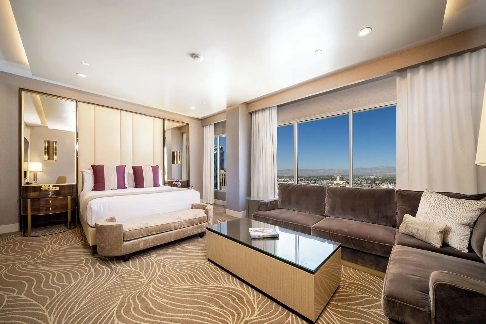 Marra Style Signature Junior Suite at SAHARA Las Vegas: a king bed with a padded headboard between tall mirrors, a velvet sectional sofa, a glass-topped coffee table and floor-to-ceiling windows