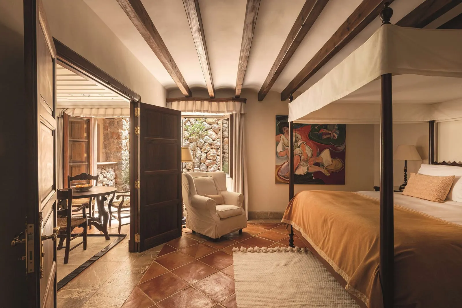 Miró Suite at Belmond La Residencia: a four-poster bed with an orange throw under wooden beams, a white armchair, a framed artwork and open doors to a table and chairs beside a stone wall
