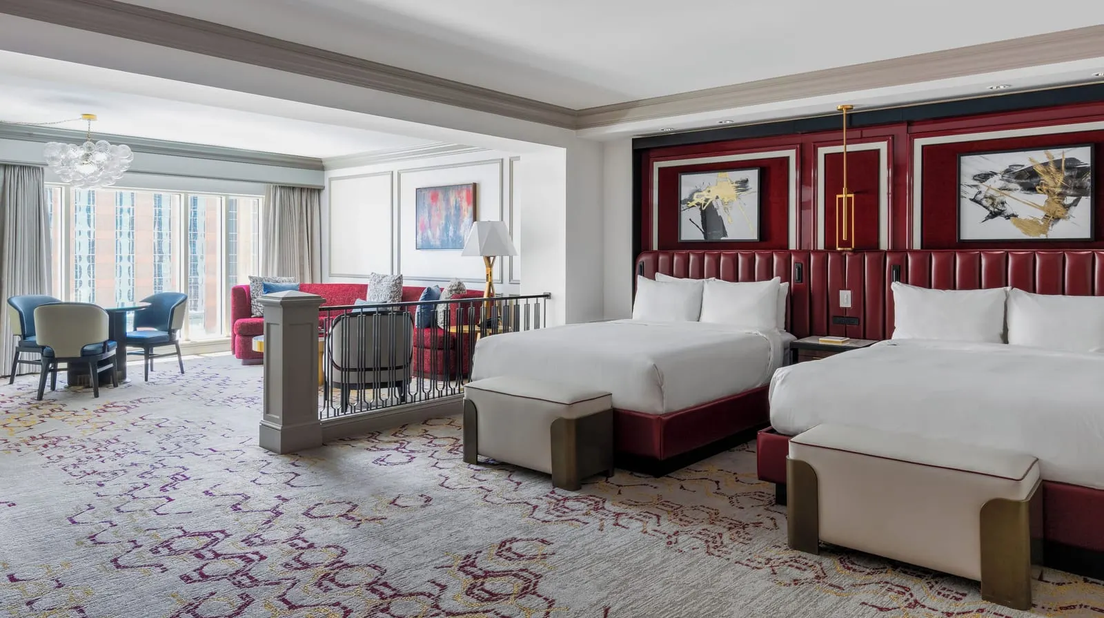 Newly Remodeled Premium Two Queen Suite at The Venetian: two queen beds against a dark red panelled wall, with a railing beside them above a living area with a red sofa, a dining table and a glass chandelier