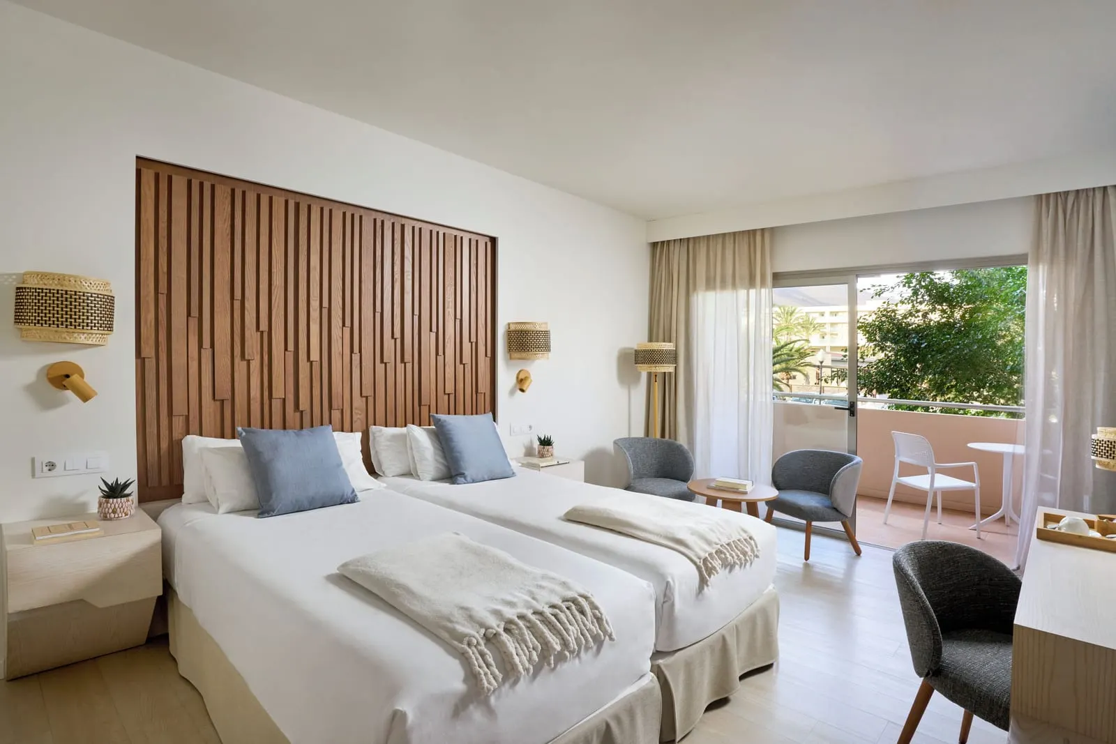 Side Sea-View Double im Iberostar Selection Fuerteventura Palace: zwei Einzelbetten unter einem Kopfteil aus Holzlatten, mit Sesseln an einer Schiebetür zu einem Balkon mit Tisch und Stuhl
