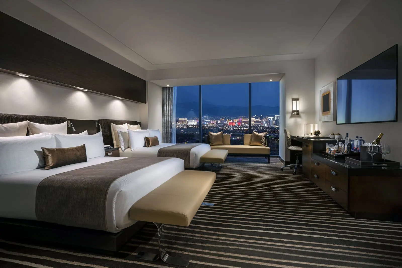 Strip View 2 Queens at Red Rock Casino Resort & Spa: two queen beds with an upholstered bench at the foot of each, a small sofa by a floor-to-ceiling window, and a long dresser below a wall-mounted television