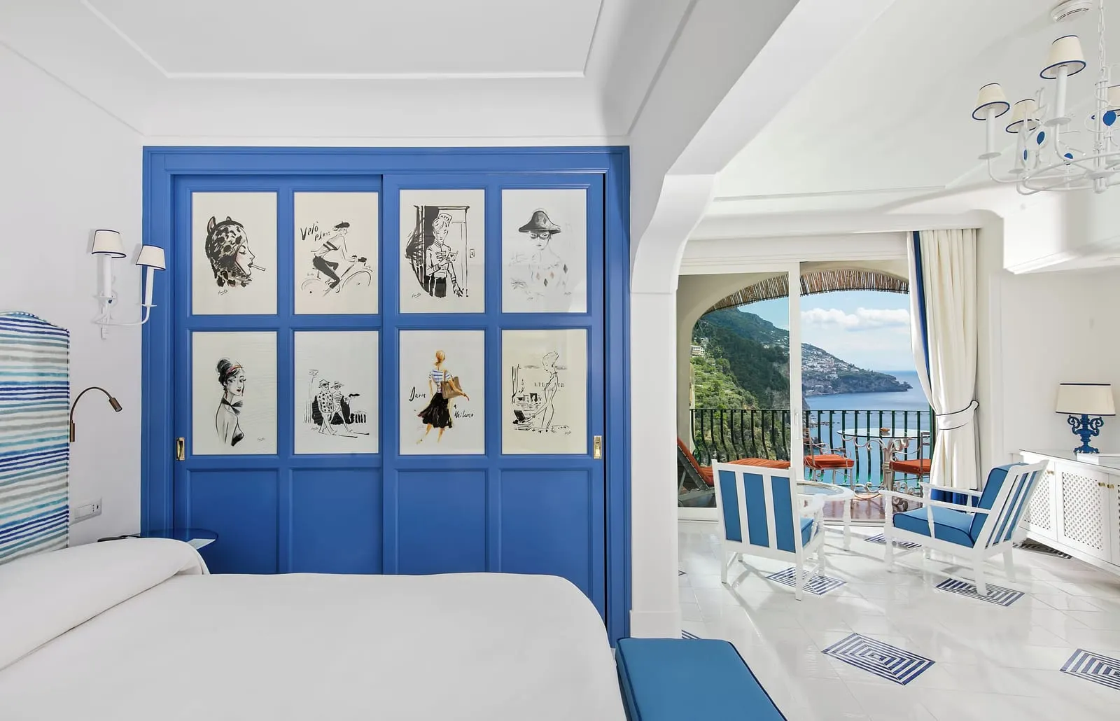 Deluxe Room at Il San Pietro di Positano: a double bed beside a blue wardrobe of framed sketches, with armchairs and a table on the terrace over the sea