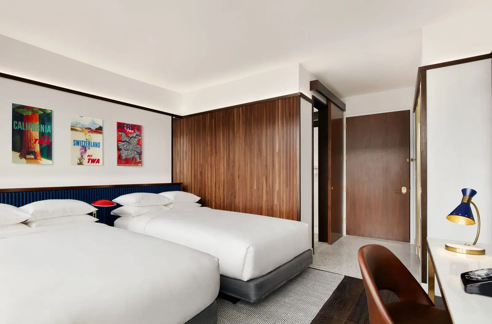 Deluxe Double Queen at TWA Hotel: two queen beds with a blue padded headboard under three framed TWA travel posters, beside a walnut slatted wall and the room door