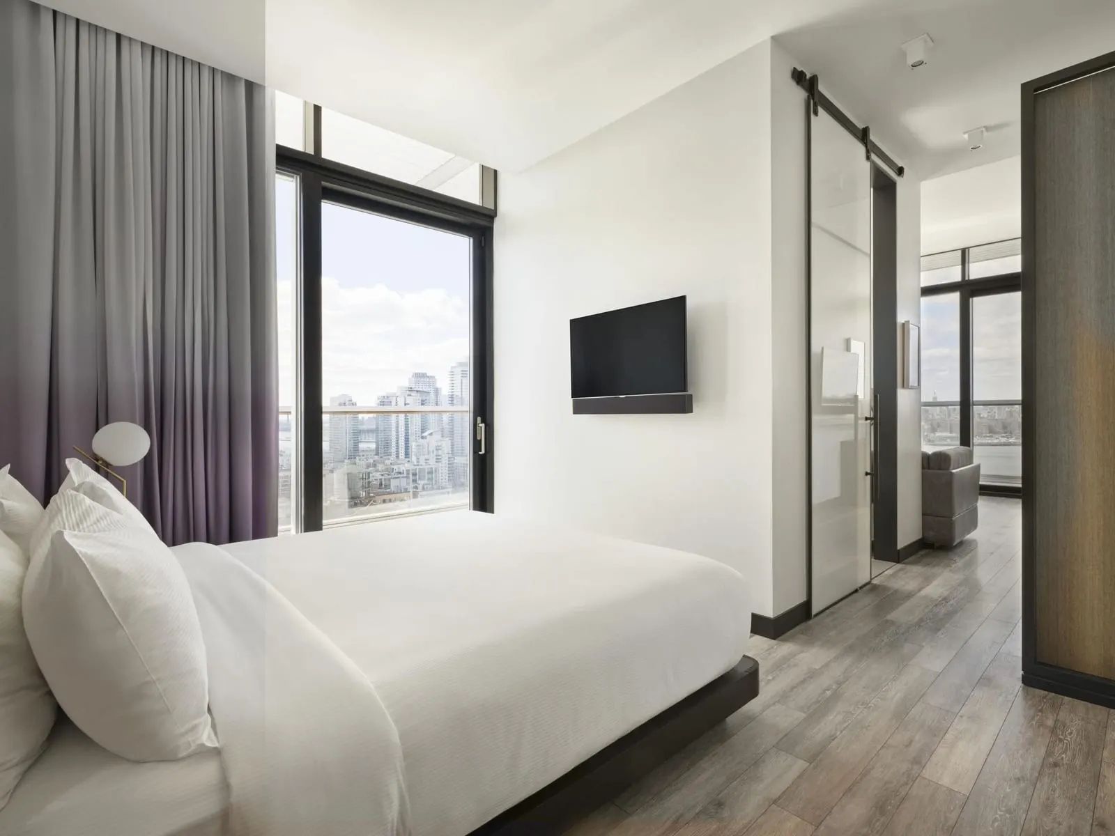 Manhattan View Corner Suite Wraparound Balcony at The William Vale: a bed with white linen beside a floor-to-ceiling window and a wall-mounted television, with a sliding glass door opening on to a sitting area with a grey sofa