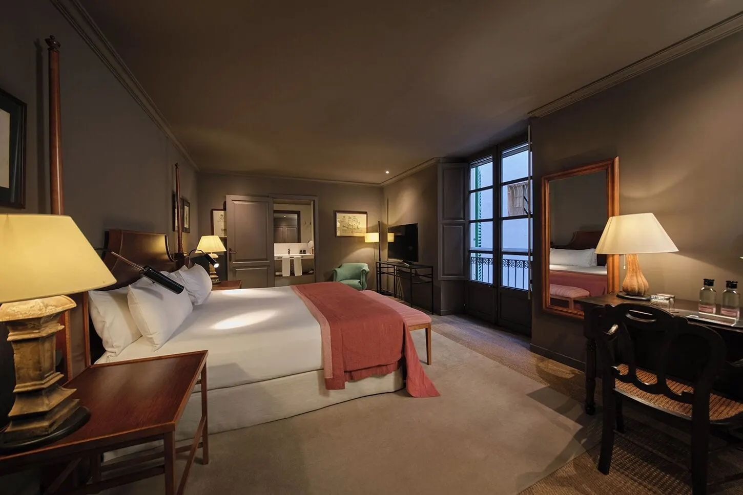 Deluxe im Can Cera: ein Bett mit einer roten Tagesdecke und einer gestreiften Bank, ein Schreibtisch mit Spiegel, ein grüner Sessel und ein offener Durchgang zum Badezimmer