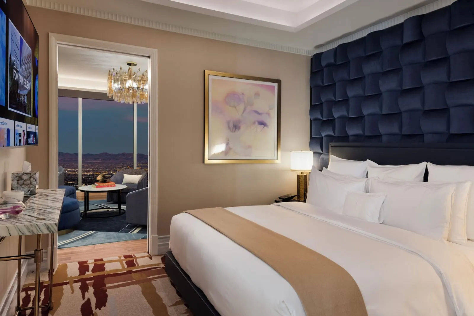 Fleur de Lis Maison Suite at Fontainebleau Las Vegas: a king bed against a wall of padded navy velvet, with a doorway through to a sitting room under a glass chandelier