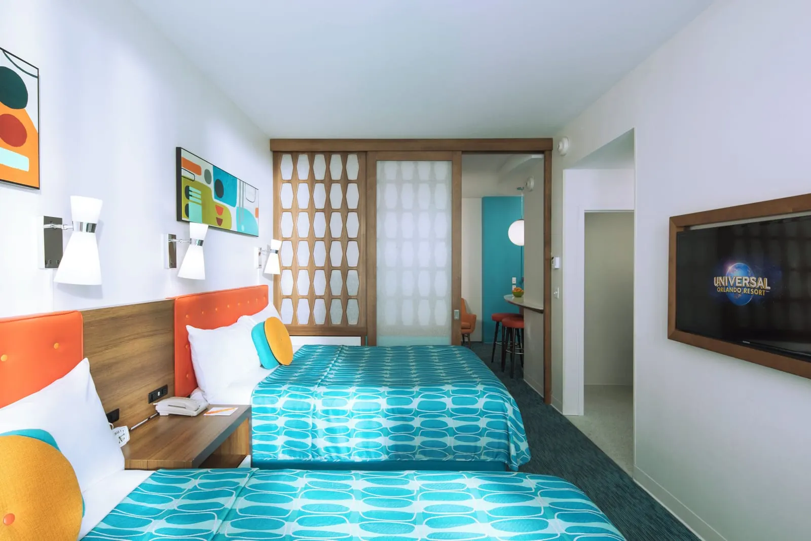 Family Suite Exterior Entry at Universal Cabana Bay Beach Resort: two queen beds with orange headboards, a wall-mounted television and the sliding lattice partition half open onto a breakfast bar with red stools