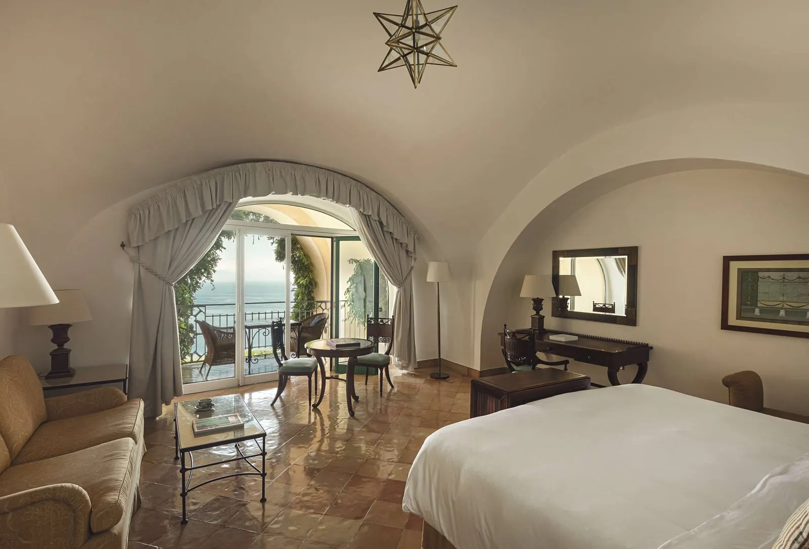 Premium Sea View Junior Suite with Terrace at Caruso, A Belmond Hotel: a bed, a sofa and a small table under a vaulted ceiling, with glass doors open onto a terrace with chairs over the sea