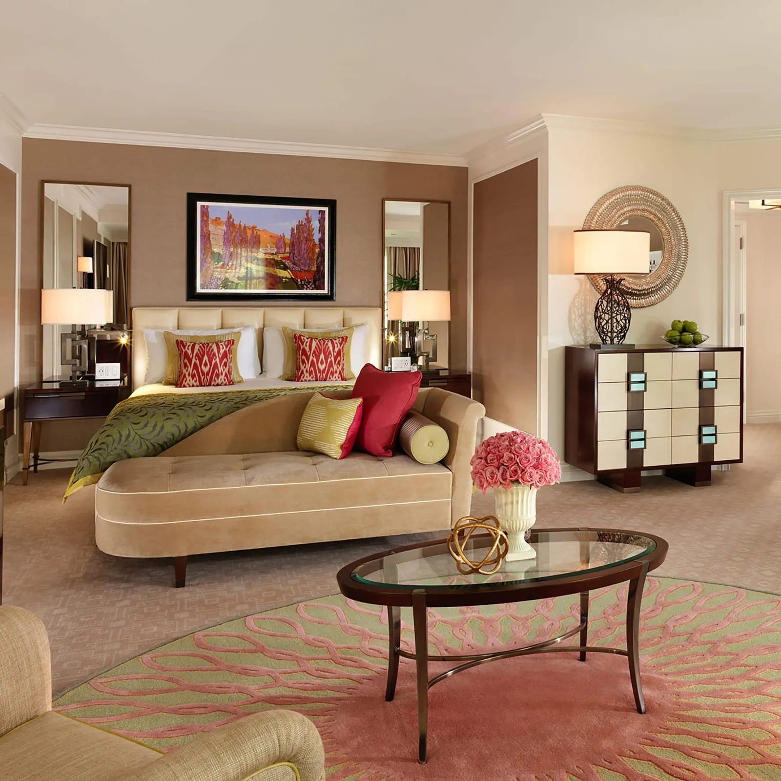 Junior Suite at The Beverly Hills Hotel: a bed with a tan chaise at its foot, a glass-topped coffee table on a round pink and green rug, an armchair and a chest of drawers, with an open door to the next room