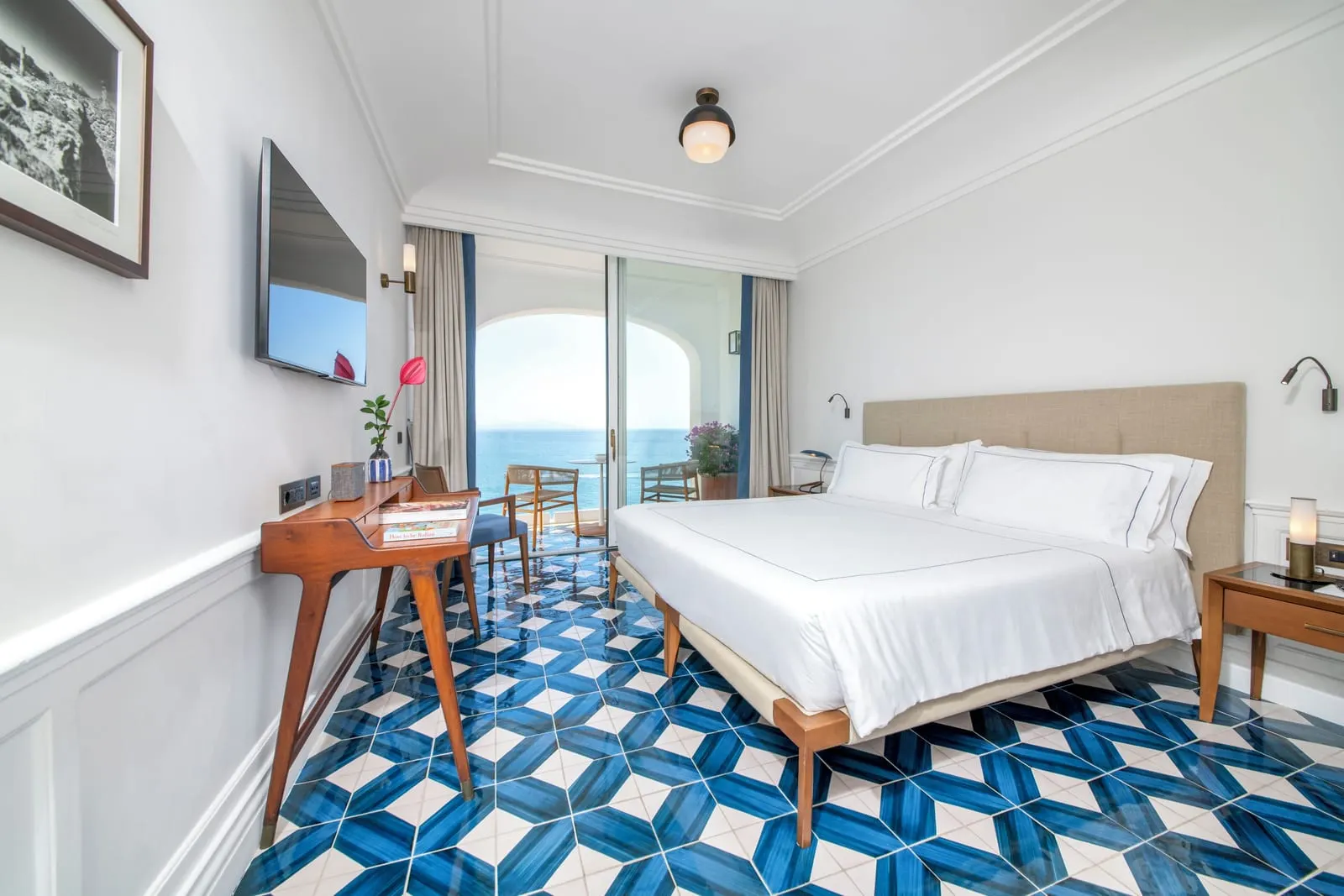 Deluxe Room Sea View & balcony at Borgo Santandrea: a king bed and a wooden desk on a blue and white tiled floor, with glass doors open to a balcony over the sea.